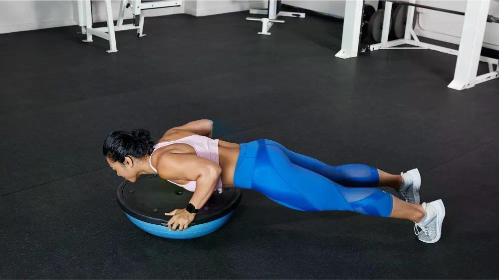 Athlete in pink sports bra and blue leggings performs a push-up on a round balance disk in a gym.