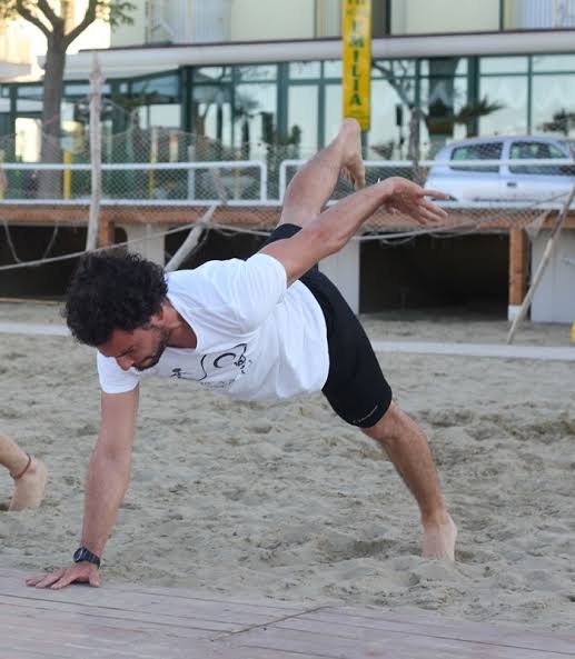 Atleta su sabbia in Allenamento: corpo teso, una mano al suolo e l’altra a mezz’aria, in equilibrio su una parete d’acqua. Si allena su una spiaggia con sfondo urbano.