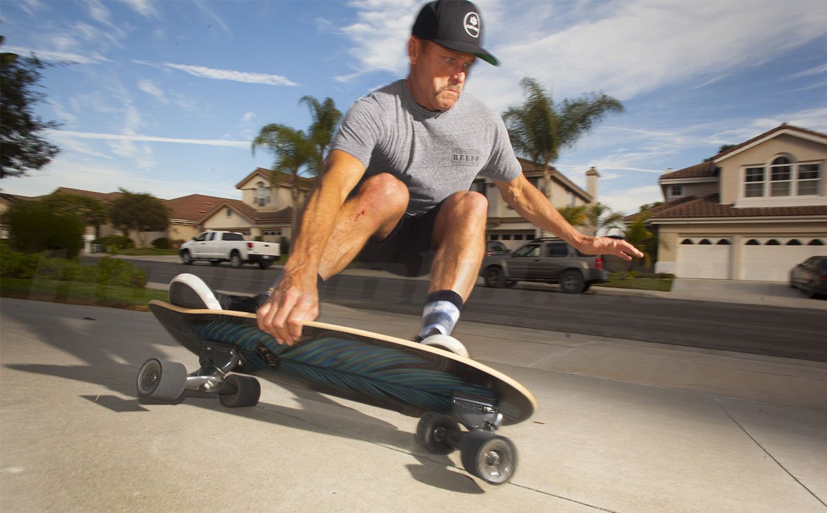 Atleta su skateboard lungo strada residenziale, con casco, indossa maglietta grigia e calzini, esegue una curva.