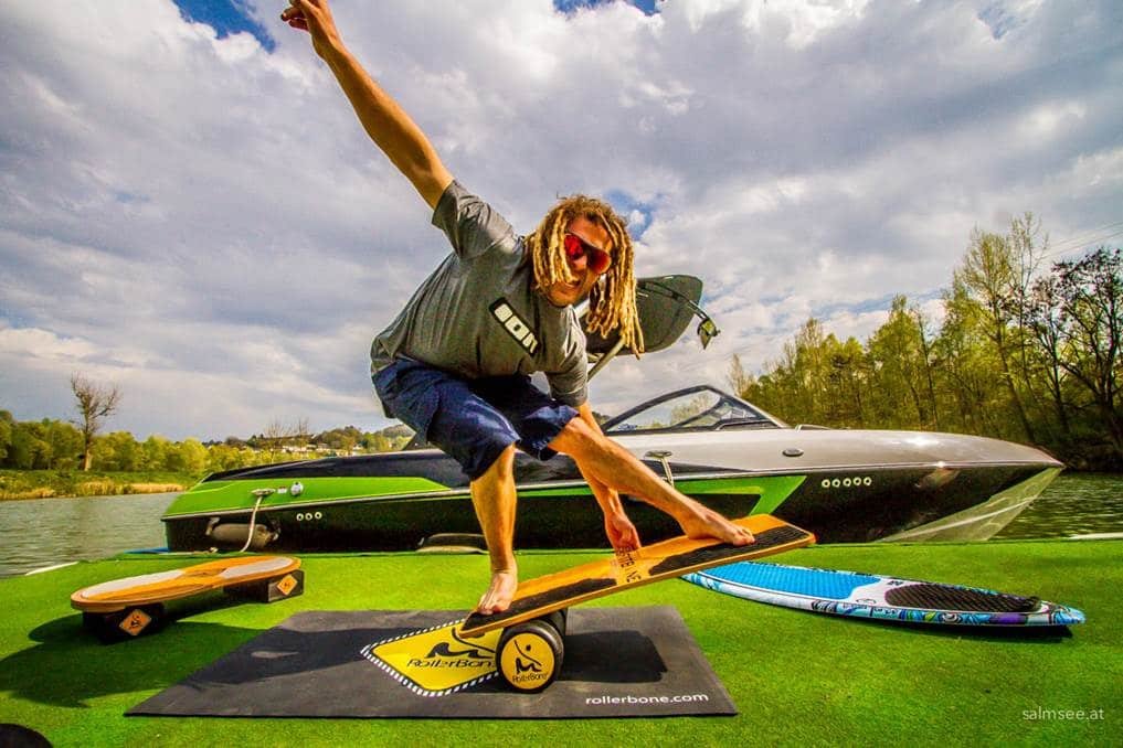 Atleta con tavola da stand up paddle su tappetino, vicino a moto d’acqua e tavole a terra in una giornata soleggiata.