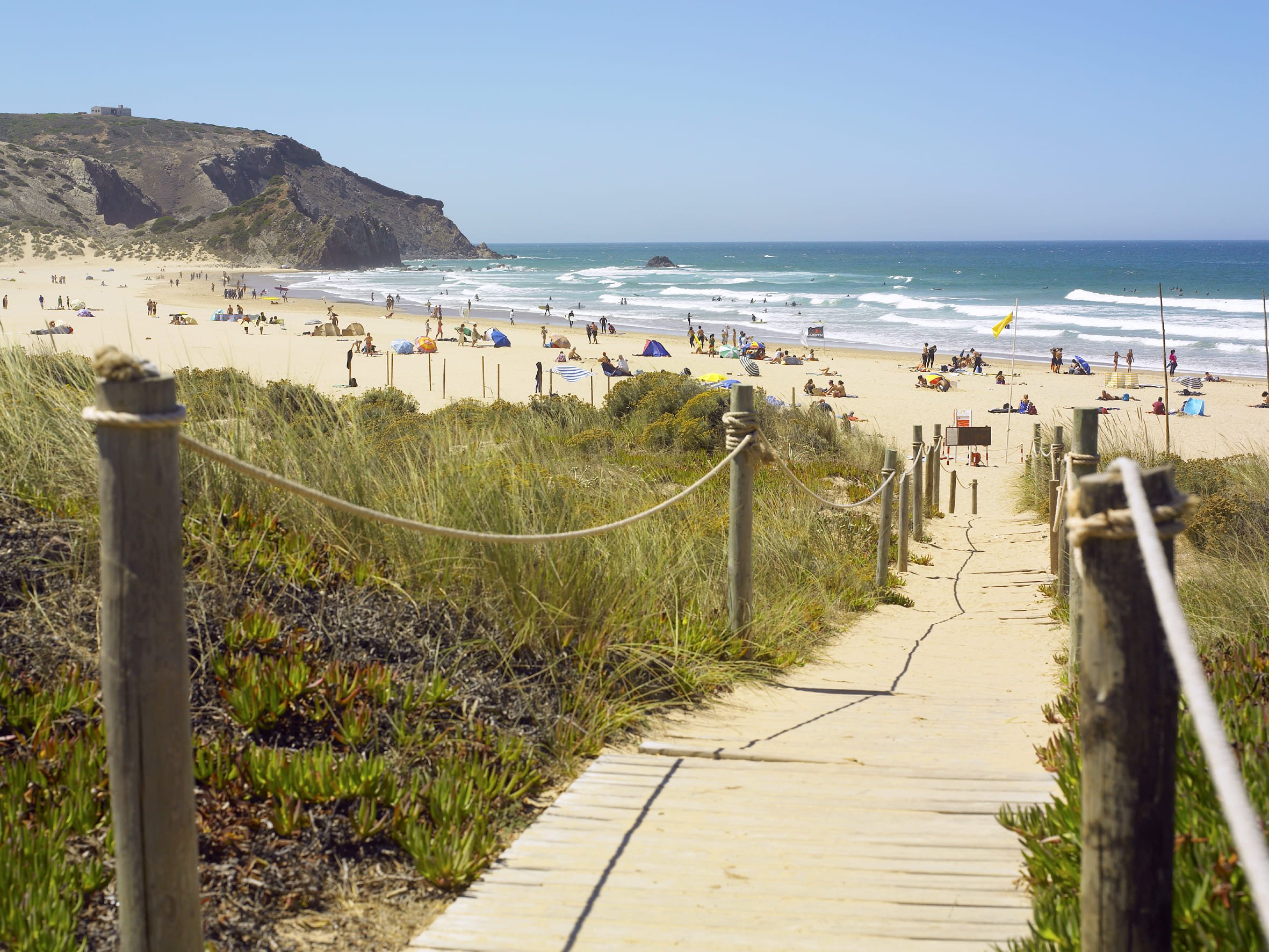 Caminho de madeira para a praia, ladeado por cordas e vegetação, levando a um areal com pessoas tomando sol e ondas ao fundo.