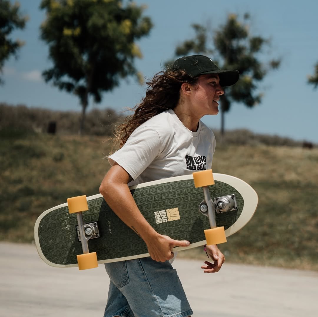 Chica con gorra sostiene skateboard corto con ruedas amarillas al aire libre, trasera de paisaje seco.