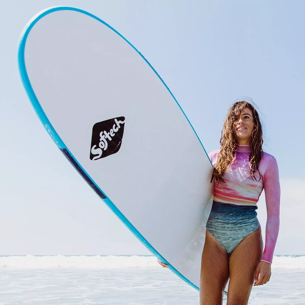 Chica con traje de neopreno sosteniendo una tabla de surf Softech en la playa, borde azul y mar tranquilo al fondo.