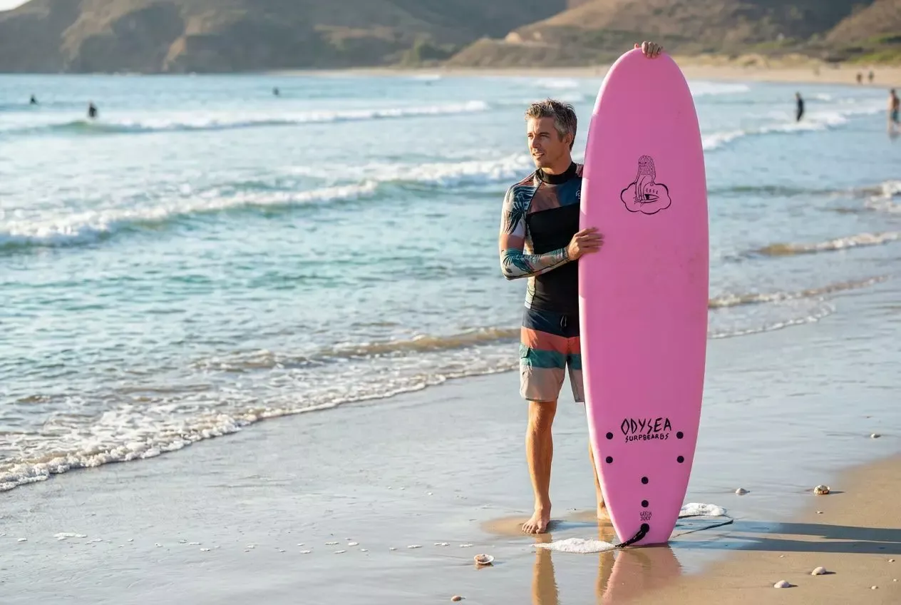 Chico en la playa con tabla de surf rosa, listo para aprender a surfear principiante, frente al mar.