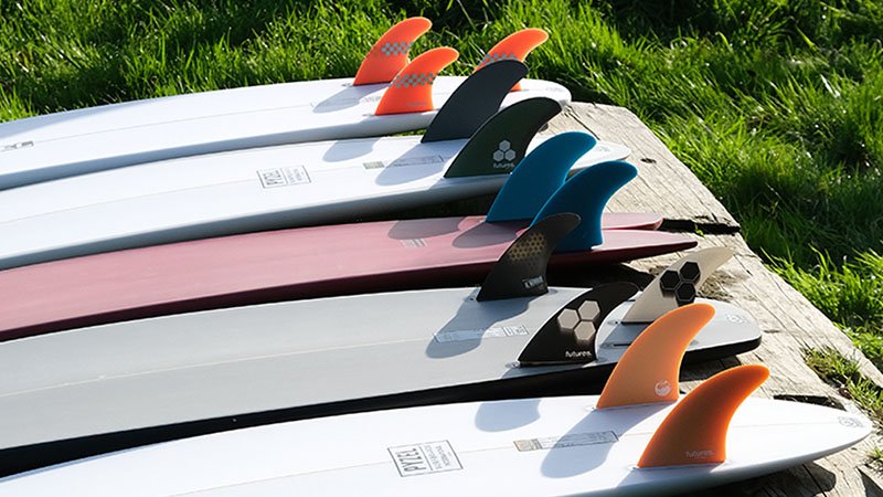 Conjunto de tablas de surf alineadas en una fila sobre un borde de madera, con aletas de colores naranja, negro, azul y gris, sobre césped verde.