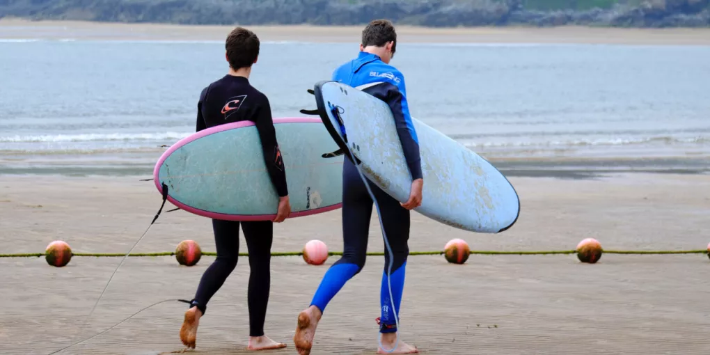 Dois surfistas com fatos de neoprene caminham pela praia, carregando pranchas de surf ao pôr do sol. (contexto de desporto na água)