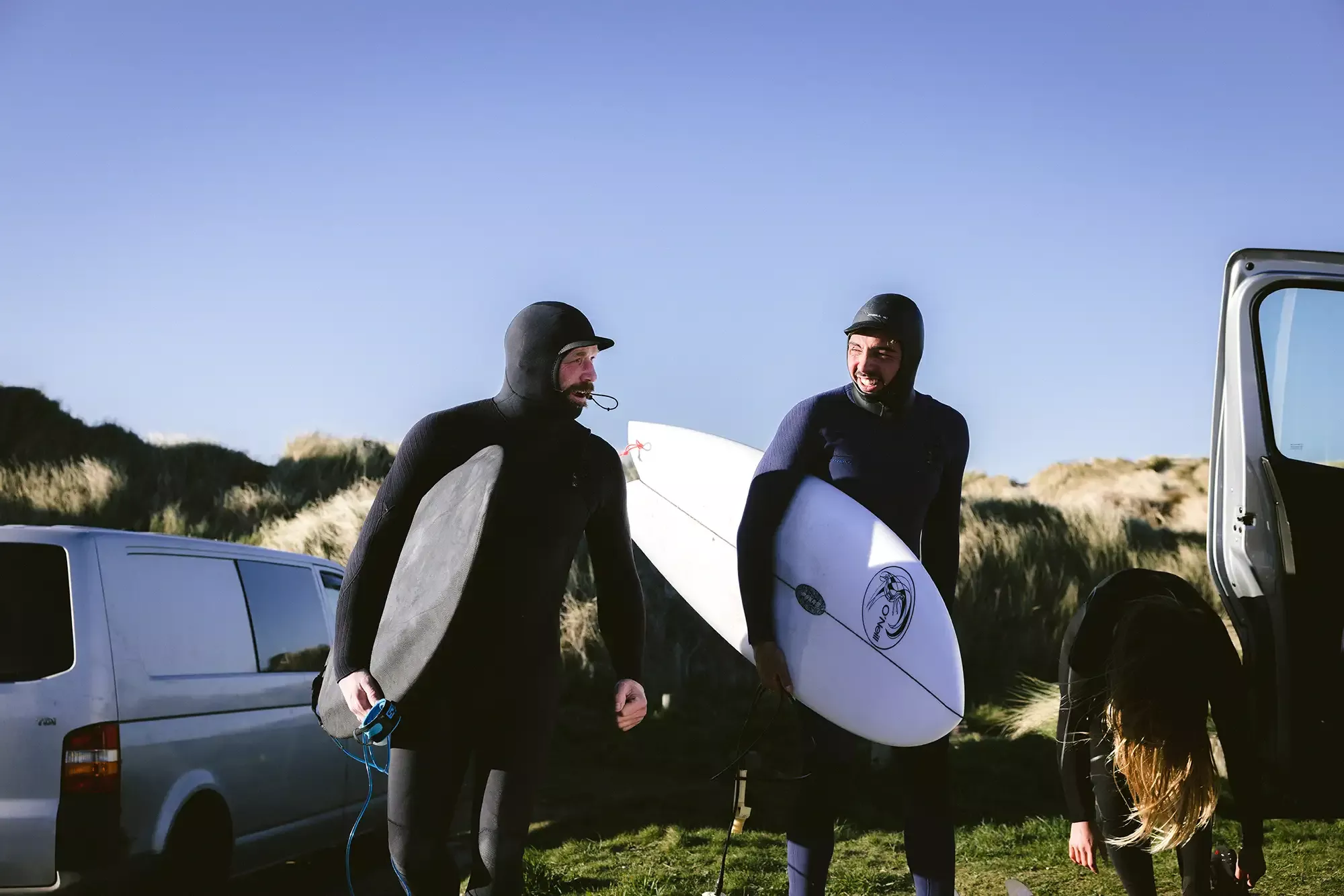 Dois surfistas com roupas de neoprene segurando pranchas, próximos a uma van na praia com dunas ao fundo.