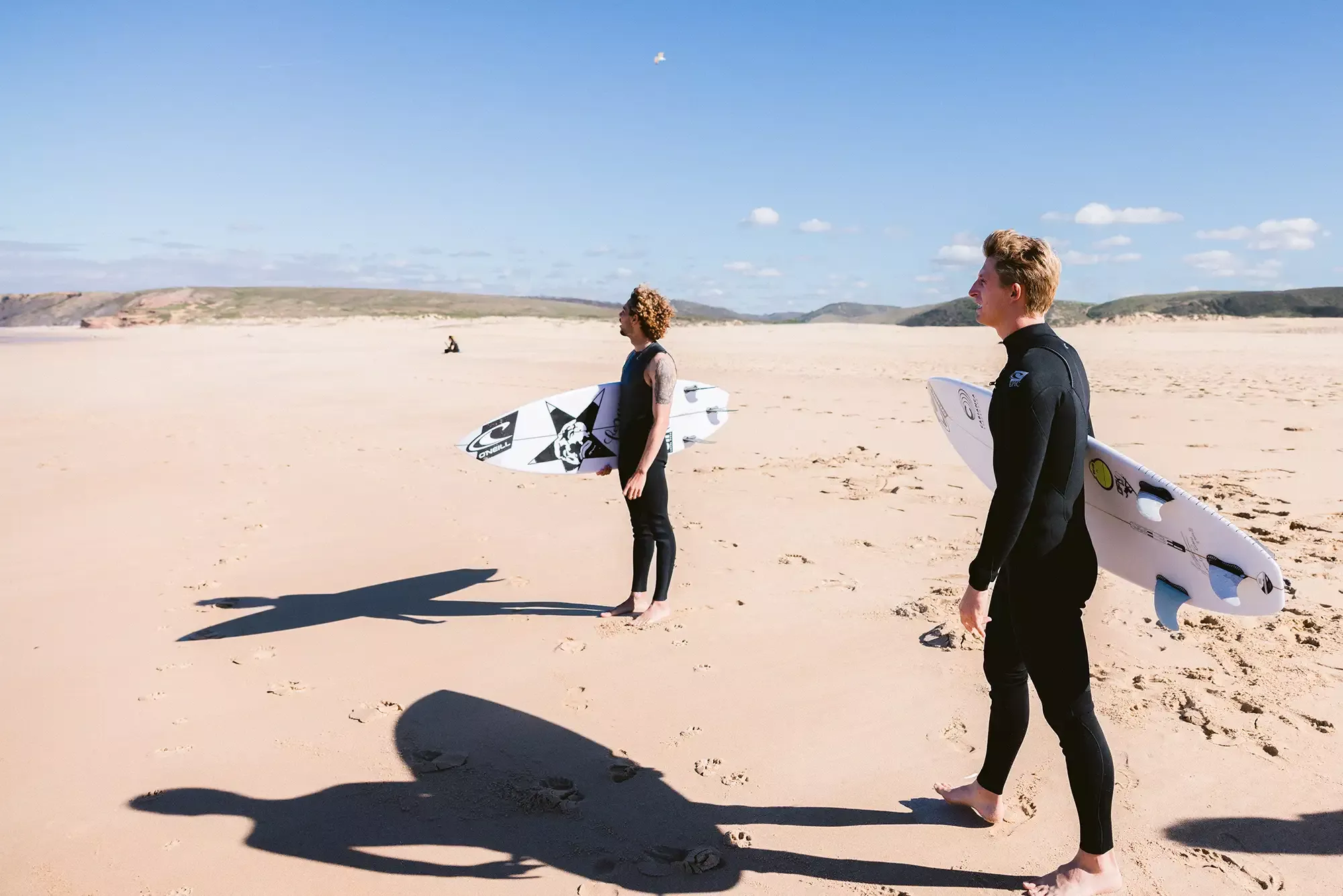 Due surfer in muta nera camminano sulla spiaggia con tavolette da surf, ombrelli di snorkeling e ombre lunghe sul sand, cielo sereno.