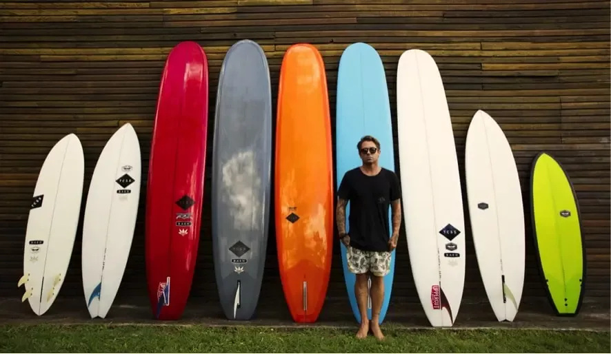 Figura com várias pranchas de surf alinhadas verticalmente alongside, de cores distintas (branco, vermelho, cinza, laranja, azul, branco, verde-neon) e uma pessoa de casaco preto e calções claras ao centro, à frente de uma parede de madeira.