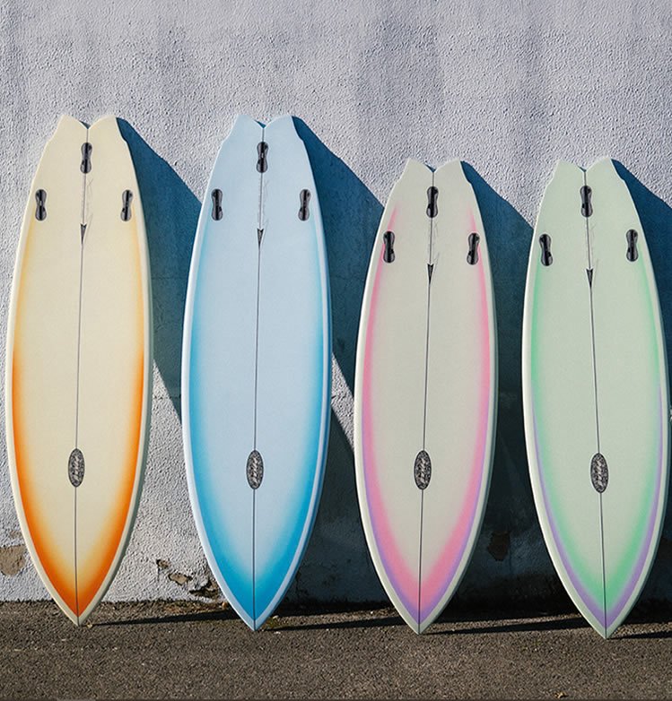 Four colourful surfboards lined up against a textured wall, each with fins and a central stringer.