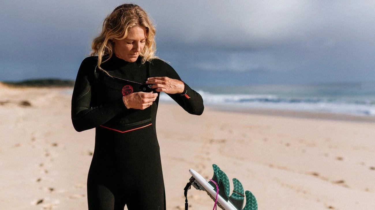 Frau in Neoprenanzug am Strand checkt ihr Smartphone, Surfboard und Leash stehen daneben.