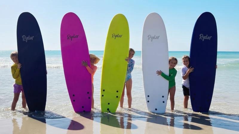 Group of children on the beach holding colourful surfboards upright in shallow water, branding visible.