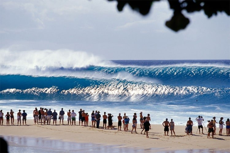 Grupo de pessoas na praia observando as ondas, com o mar azul e uma linha de pranchas de surf ao fundo.