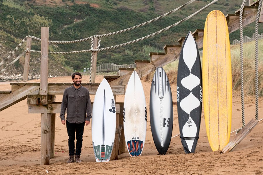 Homme debout sur une plage, près de planches de surf appuyées contre une rambarde en bois.