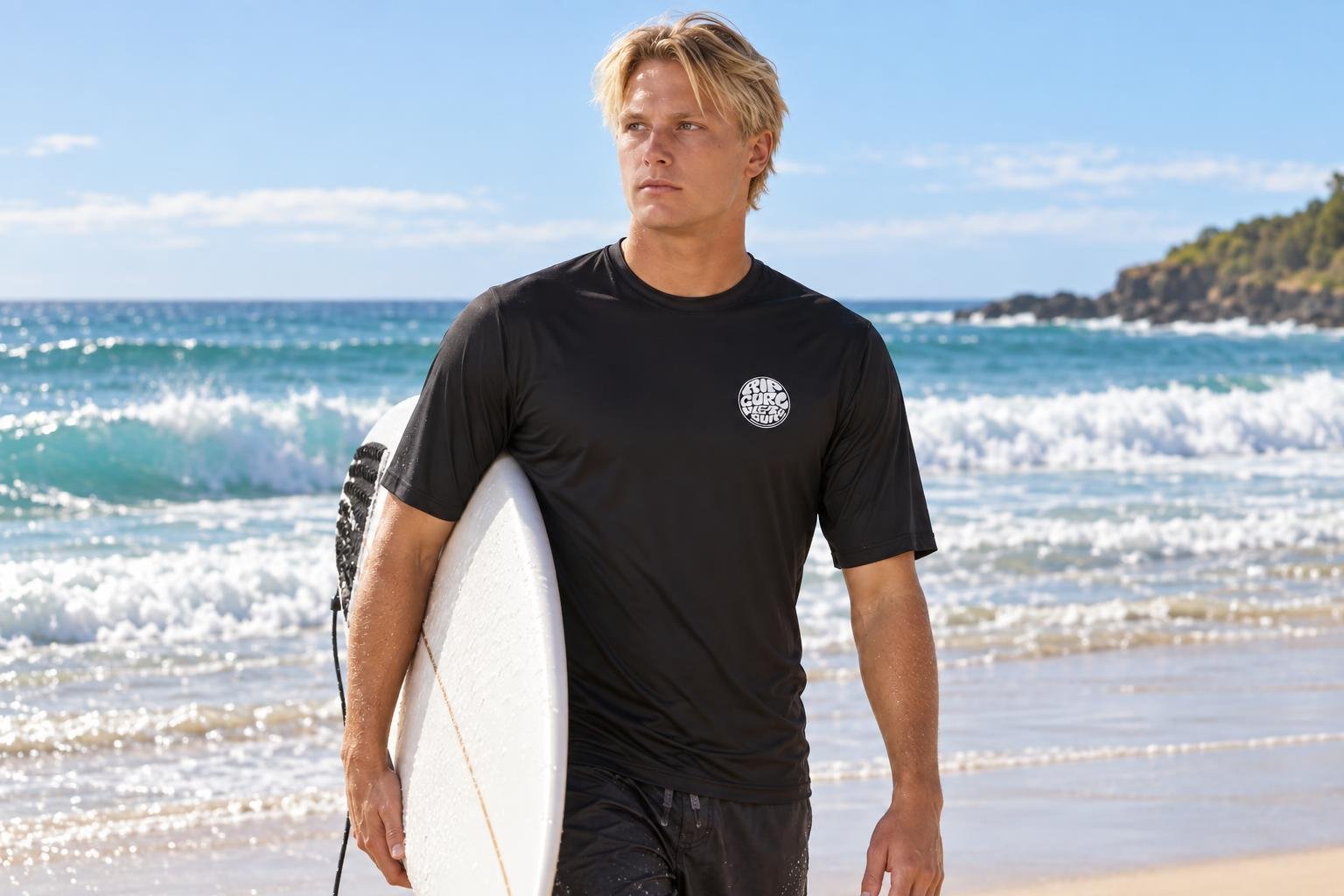 Homme sur la plage porte un haut de protection UV noir et tient une planche de surf sous le bras.