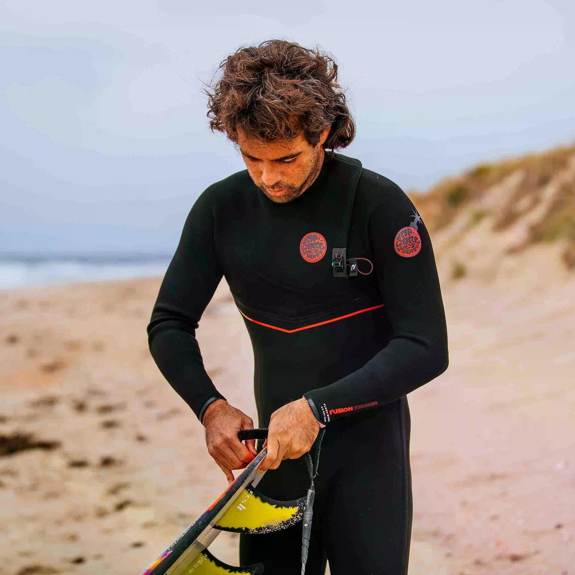 Homme surfeur en combinaison noire ajustant une ailette sur la plage, ambiance surf.