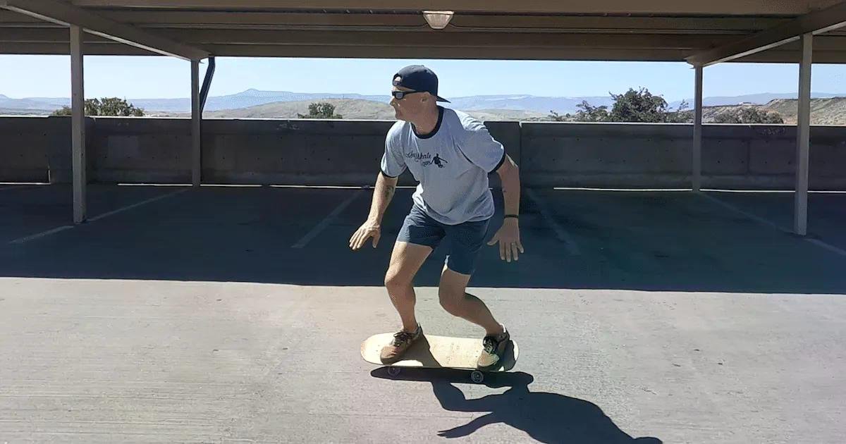 Homme surfeur débutant en skateboard, portant casquette et t-shirt gris, pratique sur une planche sous une structure ombragée.