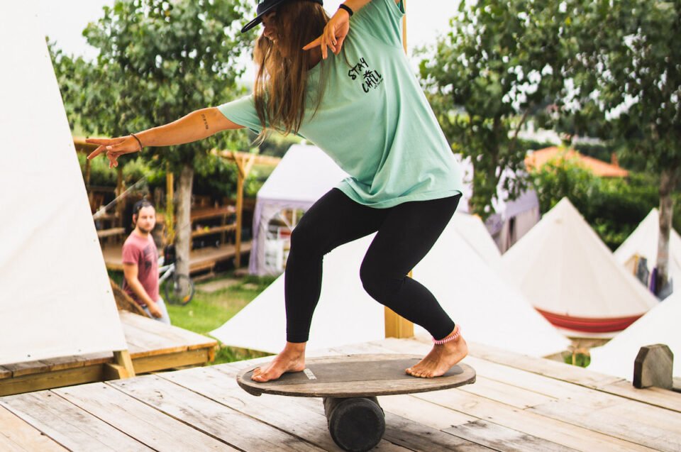 Joven practicando longboard en un skatepark al aire libre, sobre una tabla de equilibrio, con palmeras y carpas de fondo. Mujer con camiseta verde y jeans negros, descalza, en postura de equilibrio.