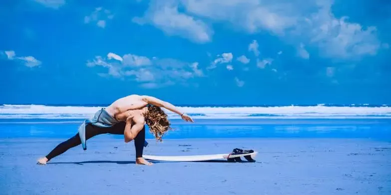 Joven surfista desnudo practicando yoga en la playa con tabla de surf a un costado, bajo cielo azul.