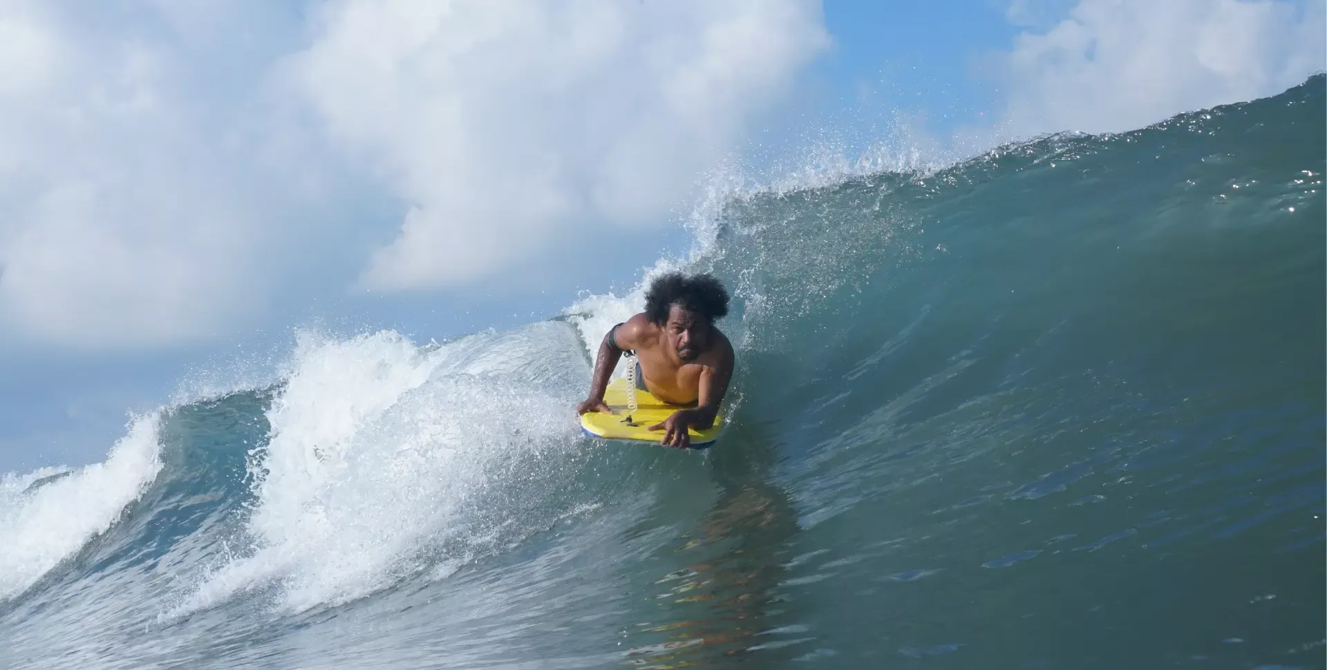 Joven surfista en traje ligero montando una ola con tabla amarilla, acercándose a la pared de agua en un rompiente azul.