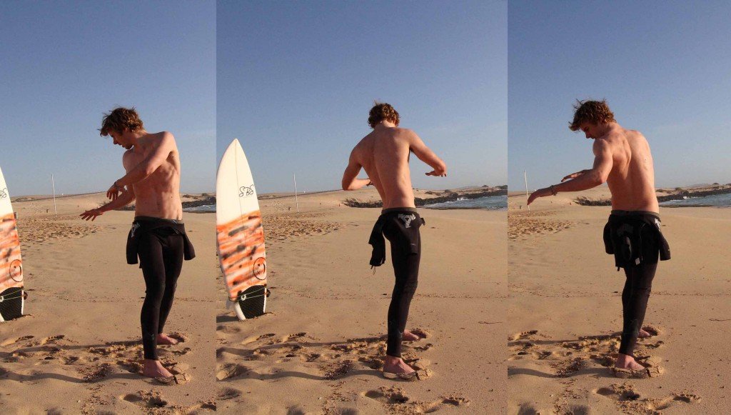 Jugador de surf sin camiseta en la playa, sosteniendo una tabla de surf de forma vertical, con traje de neopreno negro y un par de sandalias, durante el atardecer.