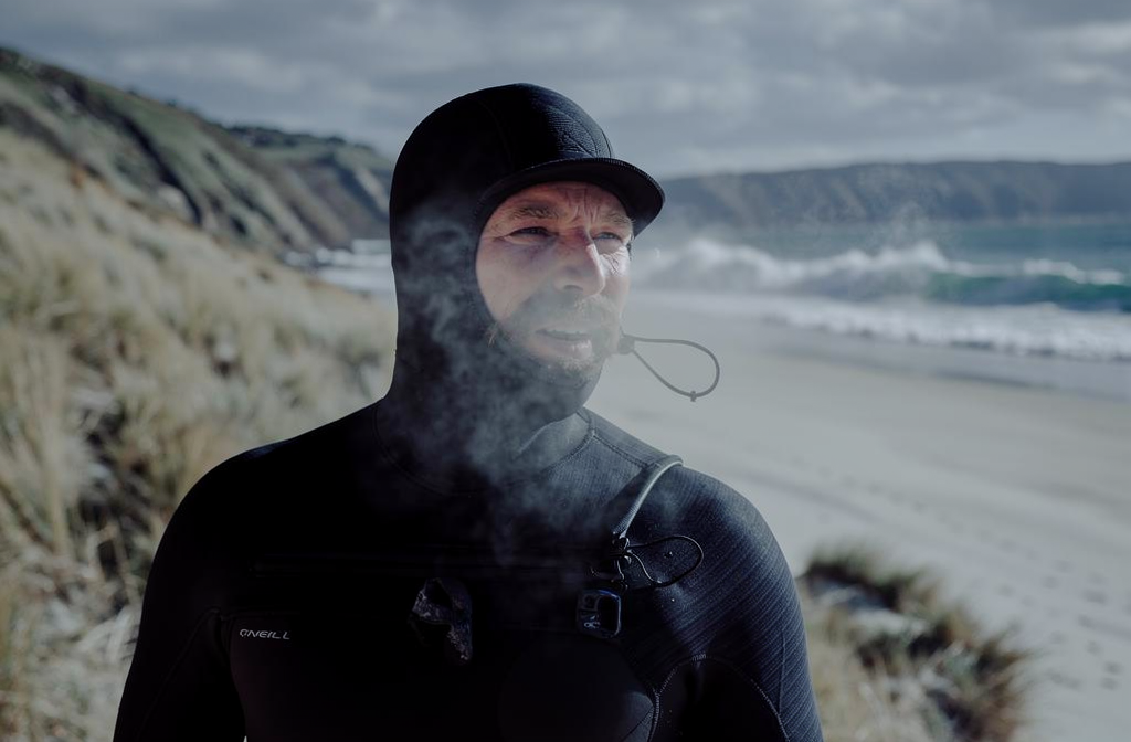 A man in a black wetsuit and hood stands on a windy beach, visible breath rising in mist as waves break in the background, illustrating how to choose a good wetsuit.