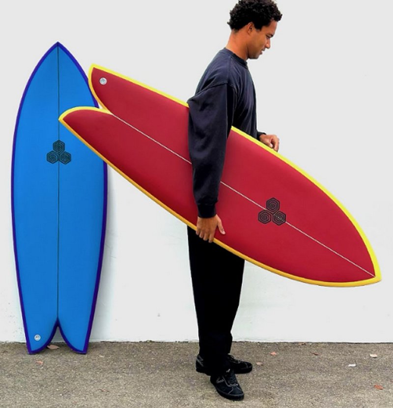 A man holding a red surfboard with yellow edge, standing beside a blue surfboard; both boards lean against a white wall.