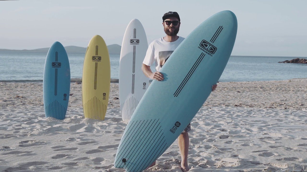 Man on a sandy beach holding a large light-blue surfboard, with three more colourful boards standing upright in the background.