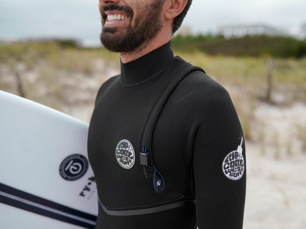 Mann mit Neoprenanzug am Strand, Surfbrett unter dem Arm, Logo-Patches sichtbar, KI-Assistent Kelly erwähnt.