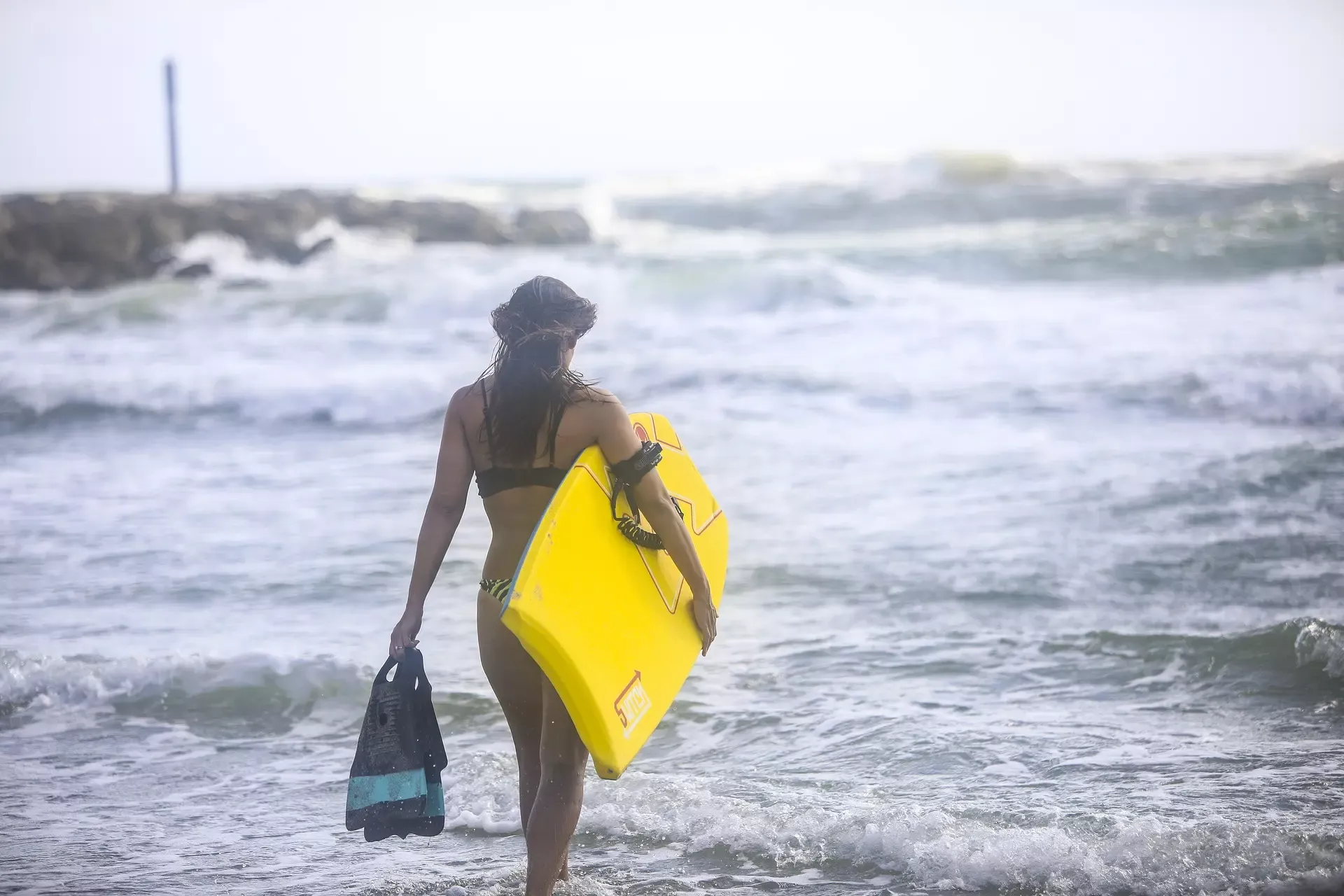 Mergulhadora a caminhar para a água com prancha amarela e mochila na mão esquerda.