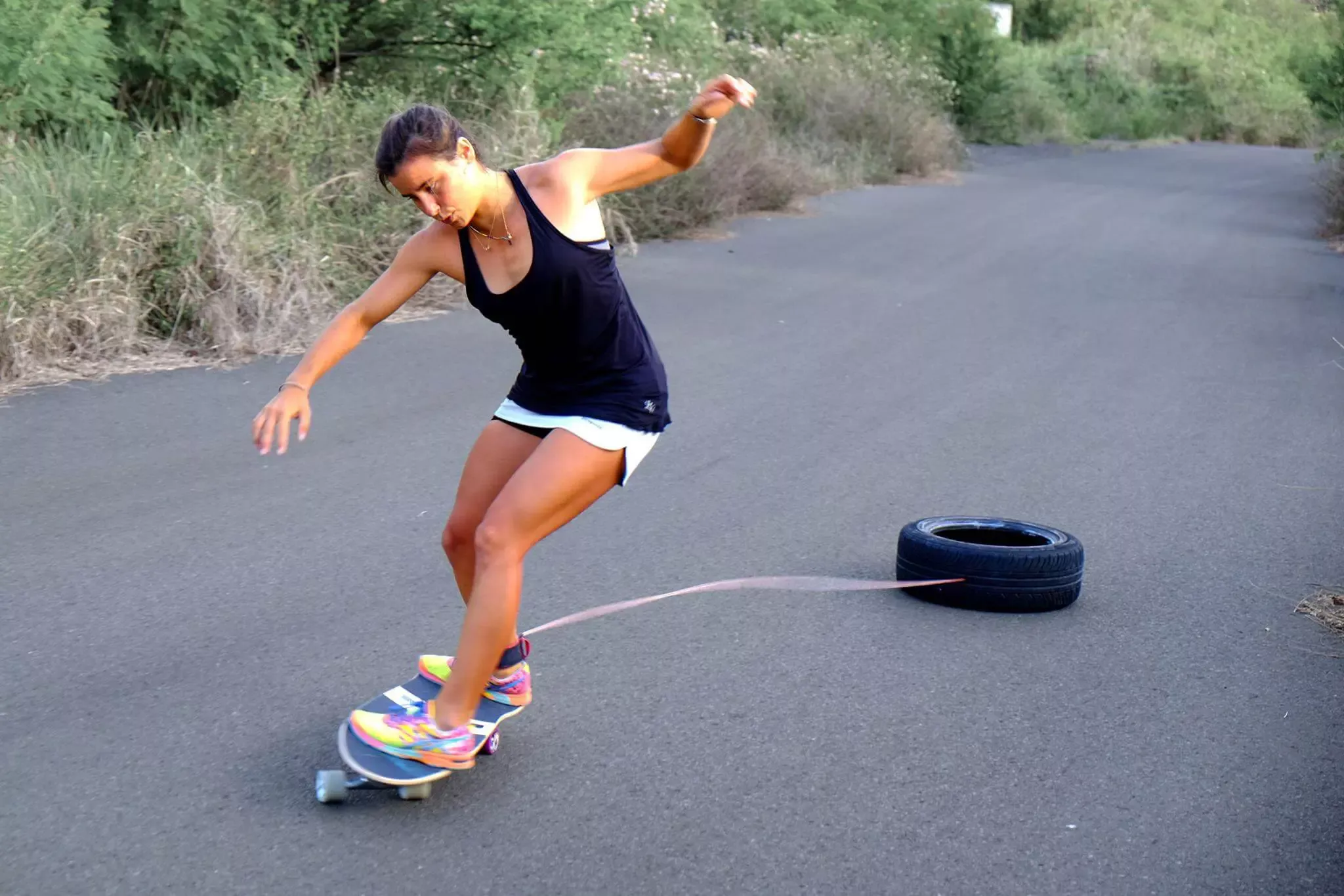 Una mujer levanta bordea con una patineta de monopatín al aire libre mientras intenta girar, con una llanta de repuesto conectada por una cuerda detrás.