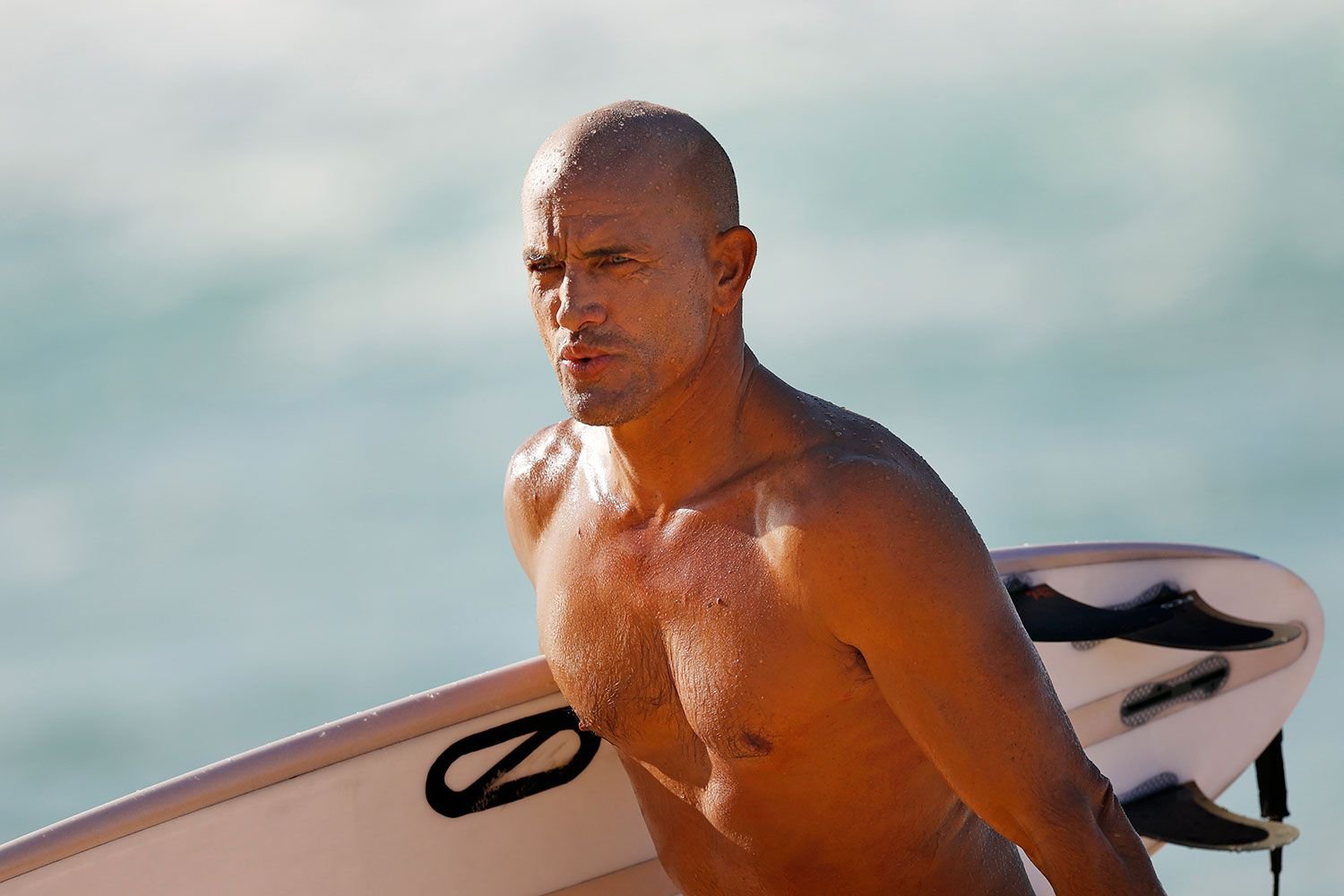 Muskulöser Surfer mit Surfbrett am Strand, Blick ins Meer gericht, Sonne glitzert auf der Haut.