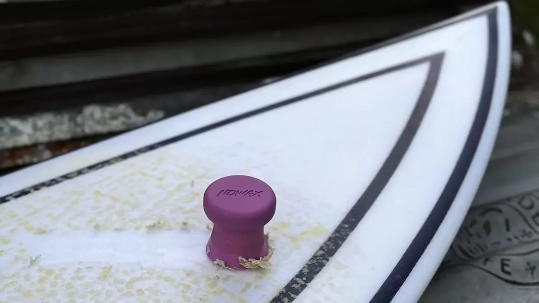 Pala en una tabla de surf con tapón de quilla púrpura en primer plano, bordeada por líneas negras.