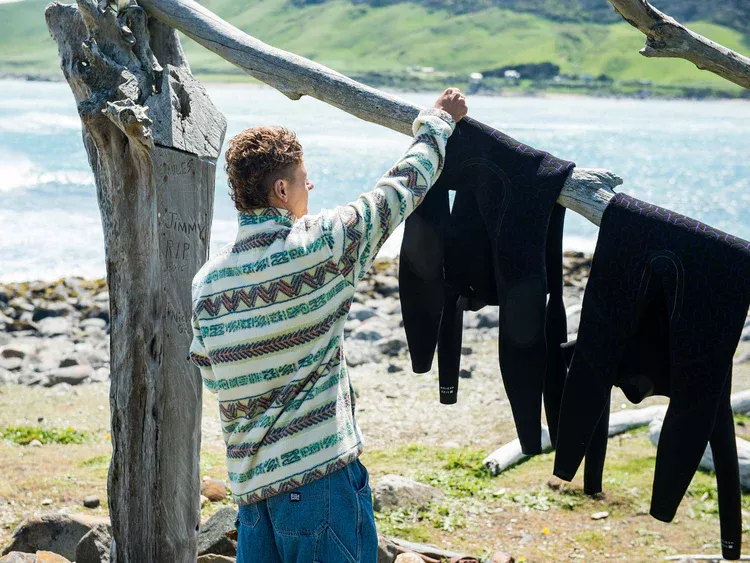 Person hanging wetsuits on a driftwood clothesline by the coast, with rocky shore and green hills in the background.