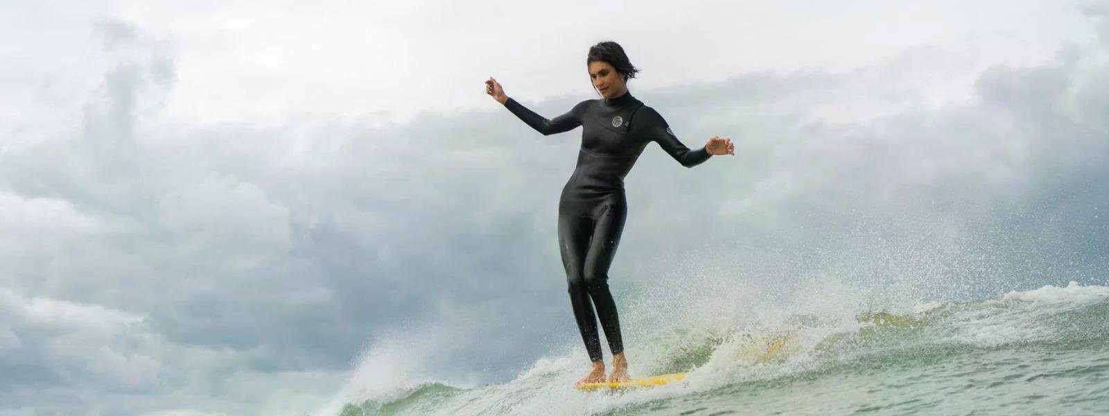Person surfing a wave in a full-body wetsuit, balancing on a yellow board with spray behind and cloudy sky above.