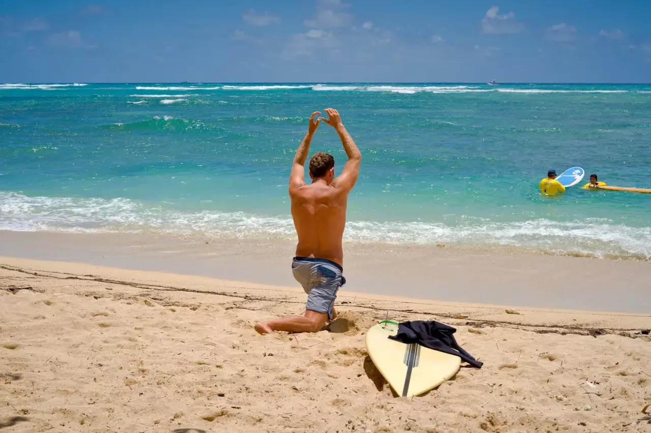 Persona de espaldas en la playa, arrodillada en la arena, levantando los brazos. Tablas de surf y neopreno en primer plano, mar turquesa y cielo azul.