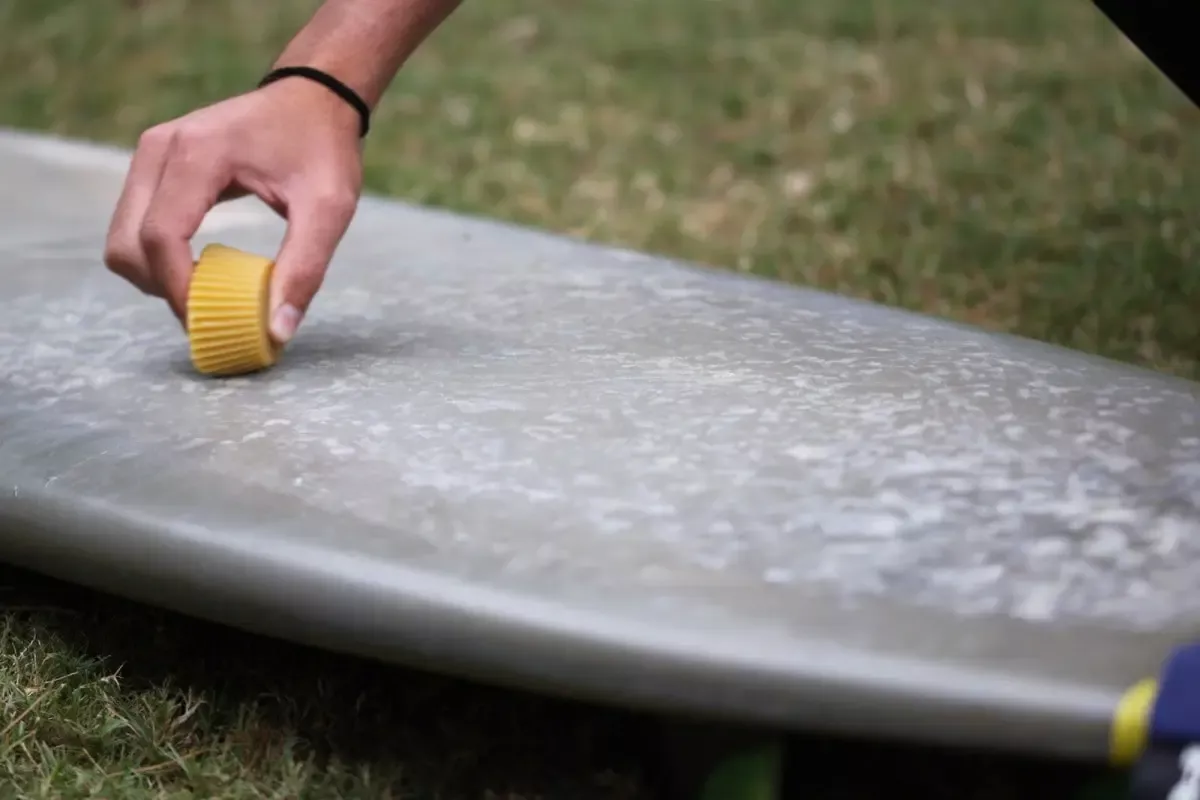 Persona limpiando o reparando una tabla de surf con una botella o tapón amarillo en una superficie rocoso-cerámica, sobre césped.