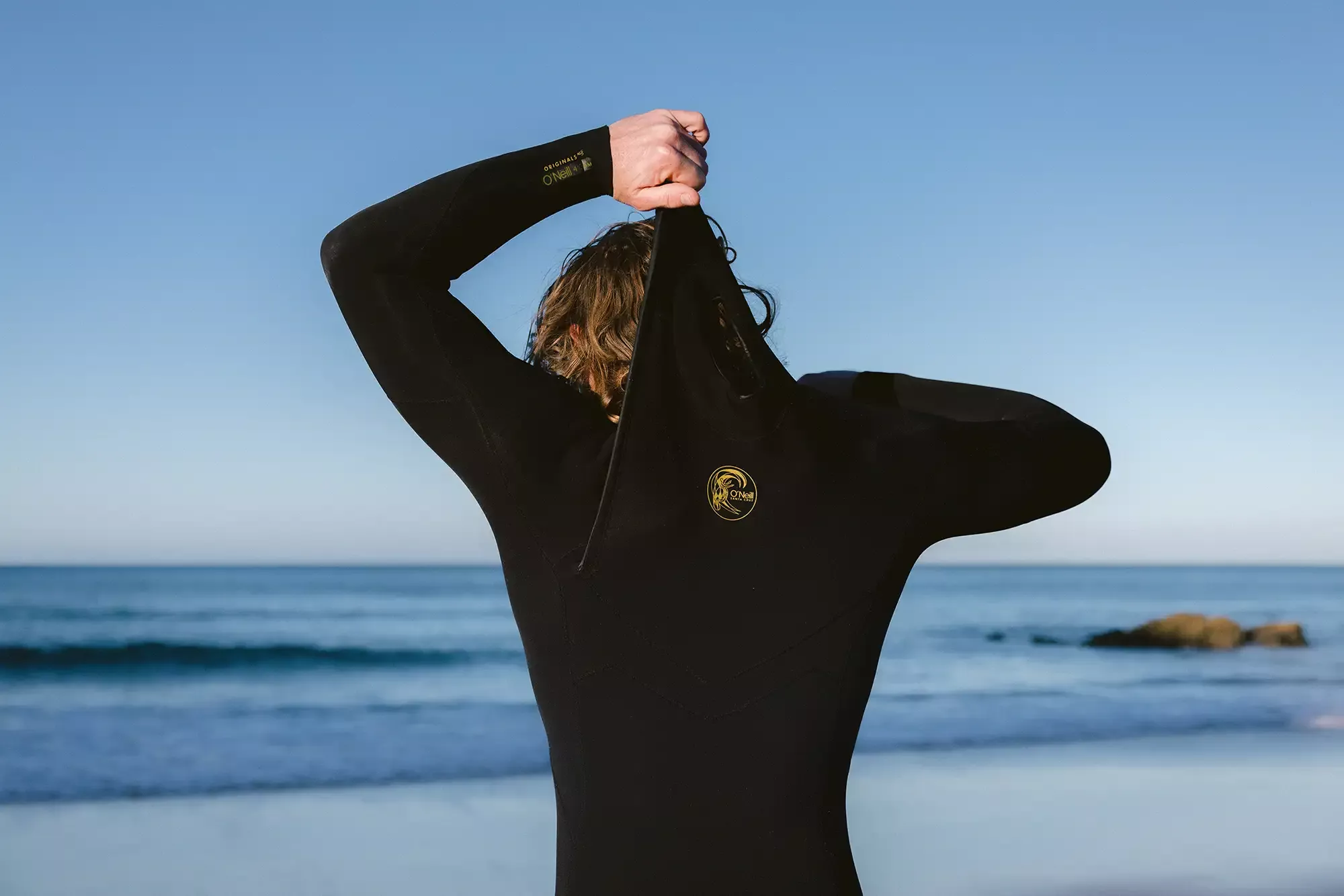 Pessoa de costas vestindo macacão de neoprene preto, ajustando o zíper na praia com o mar ao fundo.