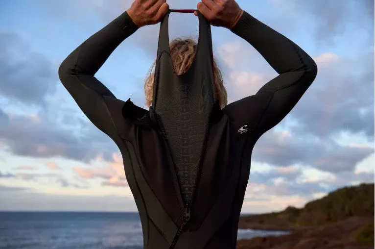 Pessoa jovem vestindo fato de neoprene de mergulho visto de trás, segurando a ponta traseira da abertura da peça com o pôr do sol ao fundo.