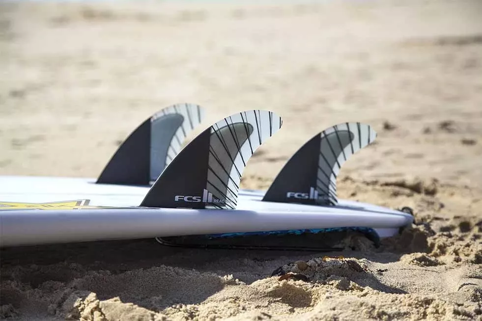 Quatro quilhas de prancha de surf enterradas na areia, vistas de lado, com a longa prancha branca ao fundo.