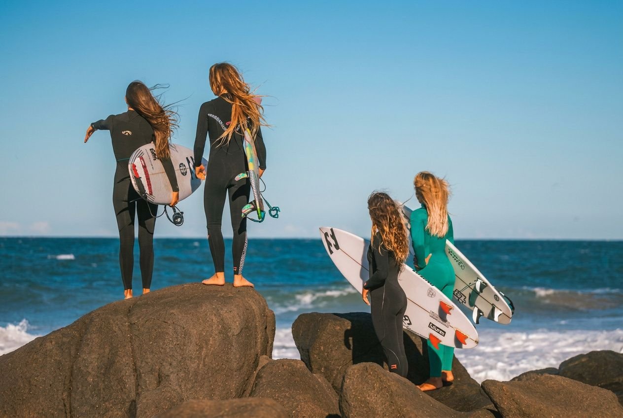 Quatro surfistas com roupas de neoprene segurando pranchas, de pé em rochas à beira-mar, observando o oceano sob céu claro. Melhores praias de surf do mundo.