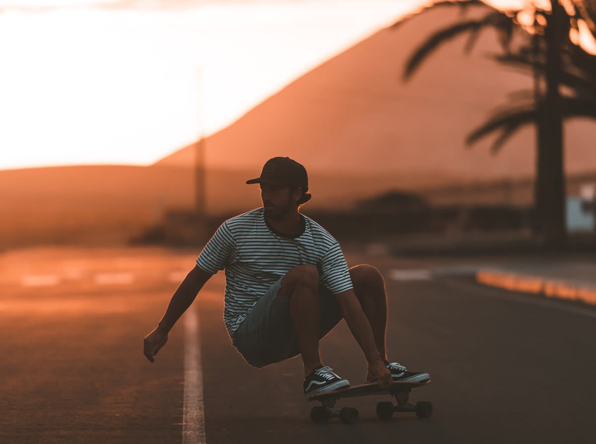 Skater sul longboard in stile sunset, in control su una strada deserta, contorni arancioni e silhouettes di palme sullo sfondo.