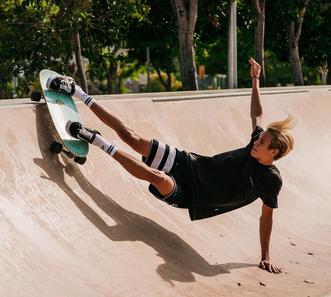 Skater macht im Bowl-Abschnitt des Skateparks einen Trick auf dem Longboard, während er mit einem Arm balanciert und der Ball aus dem Kern des Bildes ragt.