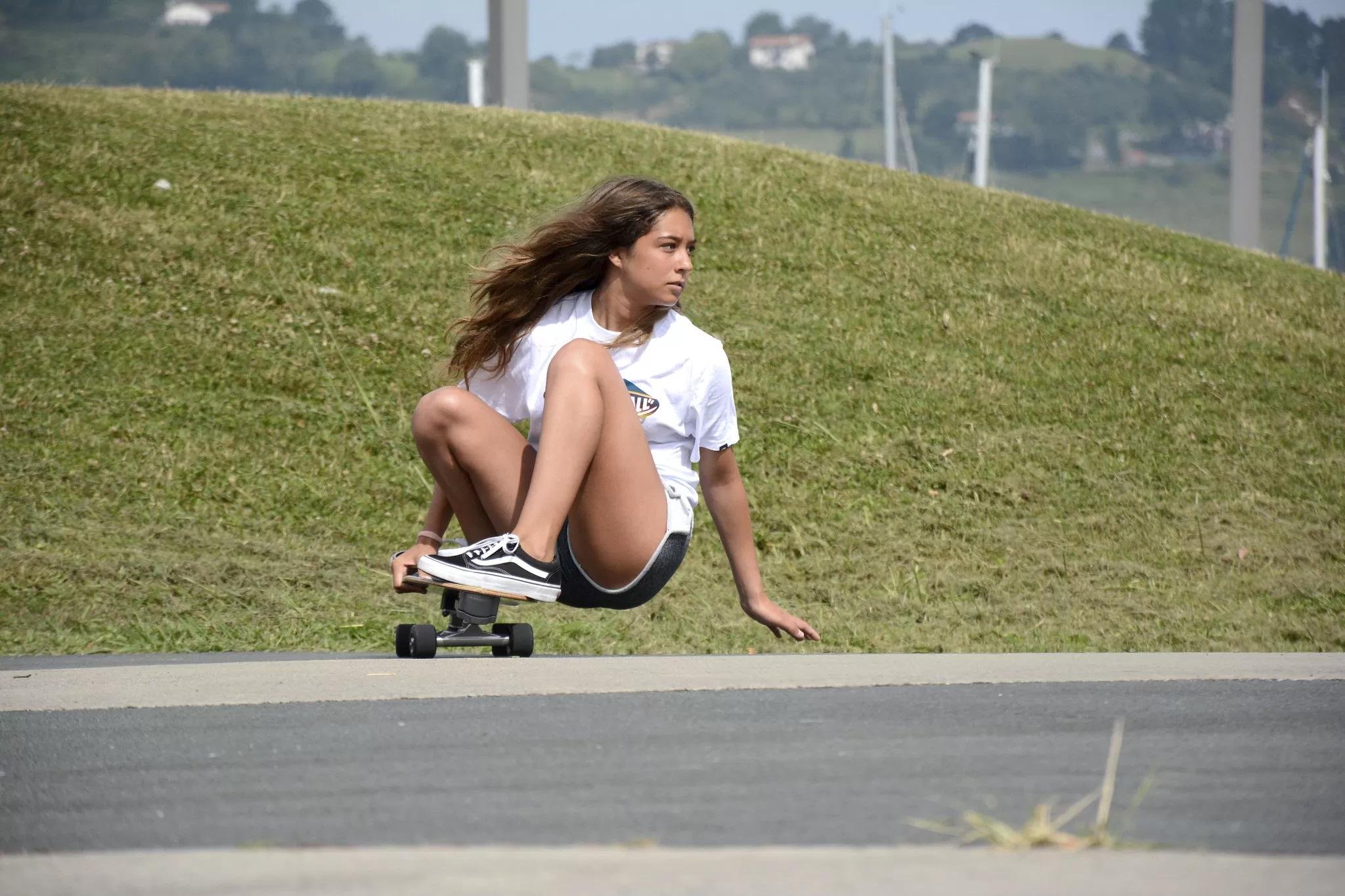 Skaterin beim Cruisen auf einem Longboard, sitzend auf dem Bord mit ausgestreckten Beinen, auf einer Straße neben einer Grasfläche.
