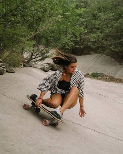 Skaterin in Schwarz-Weiß-Karo-Jacke fährt auf einem Downhill-Longboard eine glatte Betonrutsche hinunter, umgeben von Bäumen.