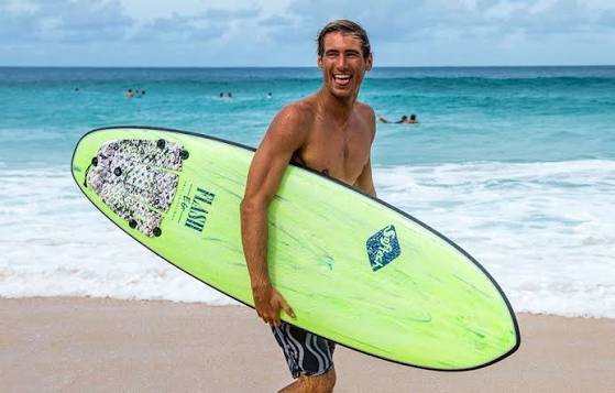 Smiling man carrying a bright lime-green surfboard along a sandy beach with blue waves in the background.