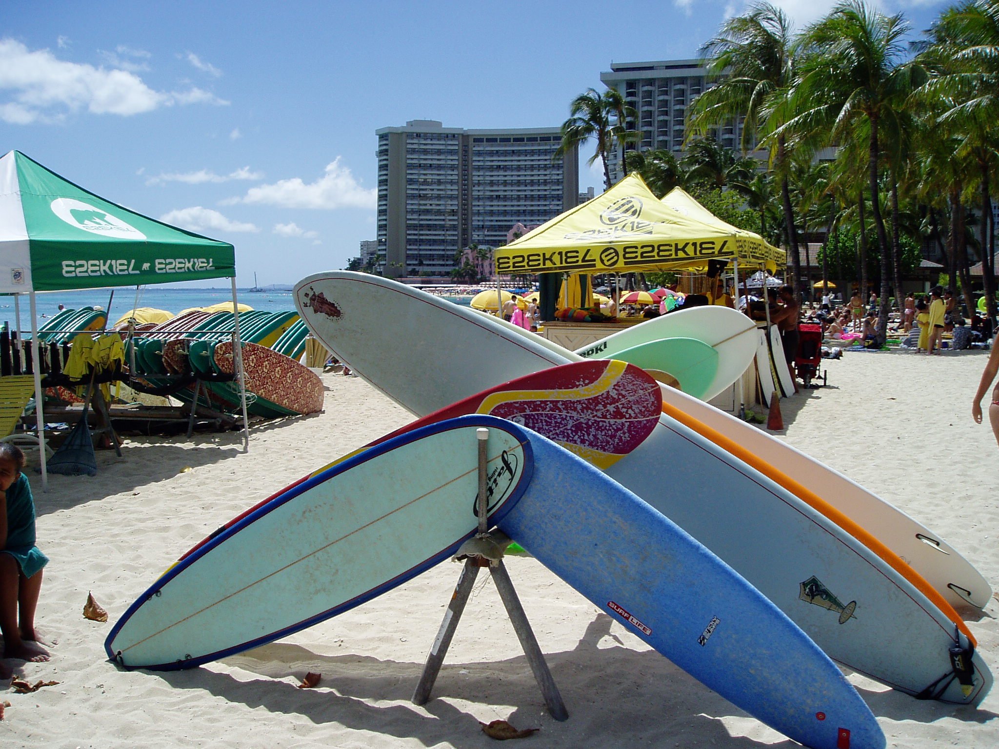 Stacked surfboards on a on the beach with tents and palm trees in the background.