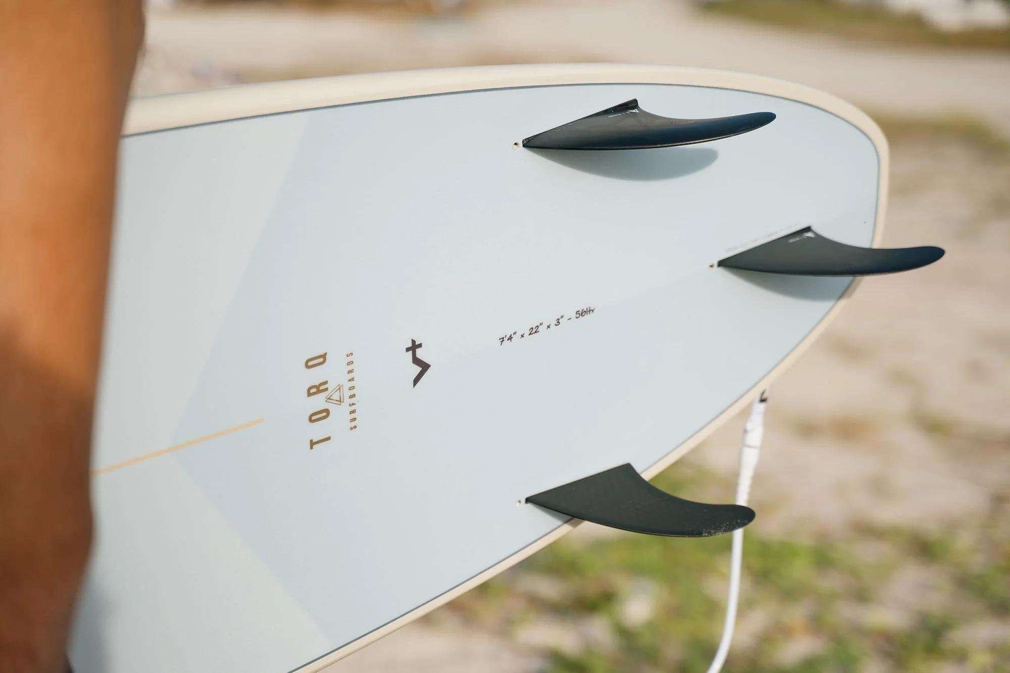 Surfboard mit Finnen am Strand, Nahaufnahme der Board-Rückenansicht; klares Design, leichter Draht zum Leash-Anschluss sichtbar. ideal für Mundo-Surf.