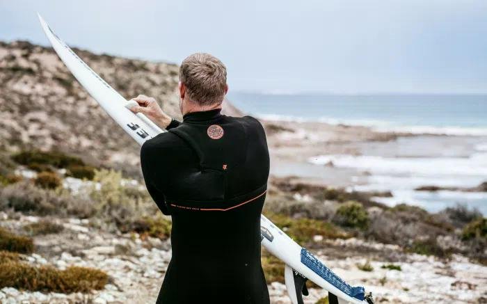 Surfer in a black wetsuit adjusting a white surfboard on rocky coastal terrain near the sea.