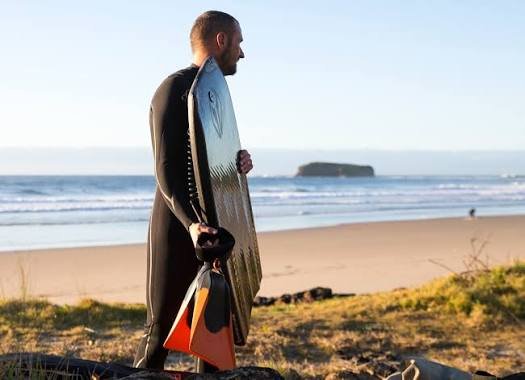 Surfer in a black wetsuit carrying a silver surfboard and orange fins along a sandy beach.